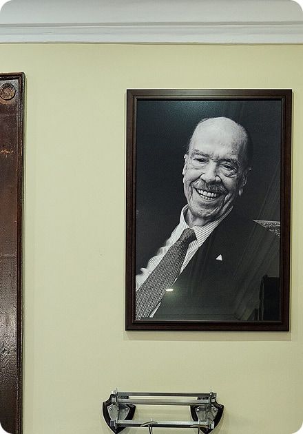 Fotografia a preto e branco, emoldurada e pendurada numa parede bege, mostrando um homem mais velho de fato e gravata a sorrir amplamente; por baixo do quadro vê-se parcialmente um suporte metálico preso à parede.