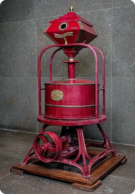 Máquina antiga de cor vermelha, em ferro fundido, com base quadrada de madeira, estrutura em arco e tambor cilíndrico central com uma placa metálica oval. Na parte superior há um funil em forma de prisma com tampa dourada e pequena abertura circular, sugerindo ser um equipamento industrial histórico, possivelmente um moinho ou torrador, exposto sobre um piso de pedra cinzenta.