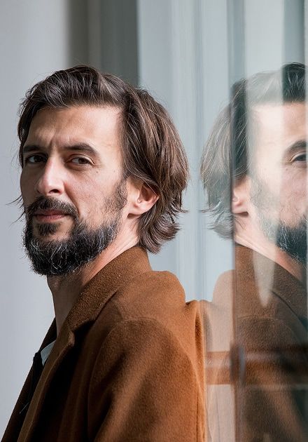 Um homem de cabelo castanho e barba, com um casaco castanho, com o seu reflexo visível numa superfície de vidro.