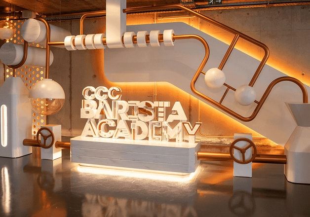 Instalação interior moderna com tubos metálicos cor de cobre iluminados por luz quente, esferas e formas geométricas brancas suspensas, e um grande letreiro tridimensional branco com a inscrição “CCC BARISTA ACADEMY” sobre uma base retangular, refletido no chão brilhante de betão polido.
