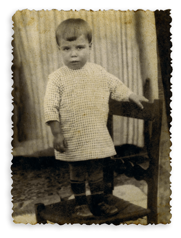 A vintage photograph of a toddler in a checkered top, standing with a hand on a wooden chair.