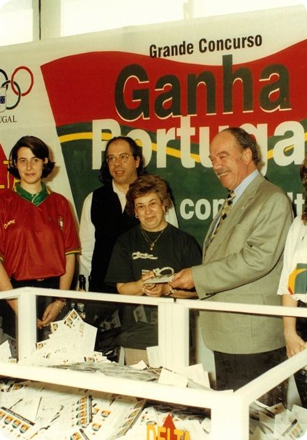 Grupo de pessoas em frente a um painel de concurso com o texto “Grande Concurso Ganha Portugal”; ao centro, um homem mais velho de fato cinzento sorri enquanto entrega um prémio a uma mulher de cabelo curto, ambos junto a uma urna transparente cheia de cupões em papel, rodeados por outras pessoas, incluindo uma jovem com camisola vermelha da seleção portuguesa.