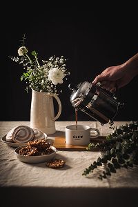 Mão a servir café de uma cafeteira francesa para uma chávena branca com a palavra “SLOW” sobre uma tábua de madeira, numa mesa coberta com toalha de linho, acompanhada por pratos com bolachas e doces, um jarro creme com flores brancas e verdes e ramos de eucalipto, sobre fundo escuro.