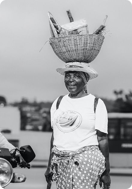 Mulher africana a sorrir, fotografada em preto e branco, caminha ao ar livre com um grande cesto de vime equilibrado na cabeça, cheio de objetos de madeira. Usa um chapéu de palha por baixo do cesto, uma t-shirt clara com estampa circular e uma capulana estampada à volta da cintura; ao fundo vê‑se um cenário urbano desfocado.