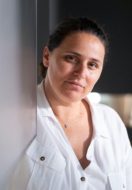 Retrato de uma mulher de meia-idade, de cabelo escuro preso para trás, encostada a uma superfície clara e a olhar diretamente para a câmara com expressão serena. Veste uma camisa branca larga com botões metálicos e um colar fino com um pequeno pendente. O fundo é interior, desfocado e escuro, com alguns pontos de luz suaves.
